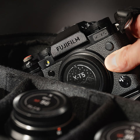 Fujifilm X-T5 avec une étiquette discrète, posé dans un sac photo rembourré, une main s’approchant pour le saisir